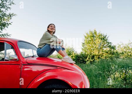 Smiling young woman hugging knees sitting on car hood Stock Photo