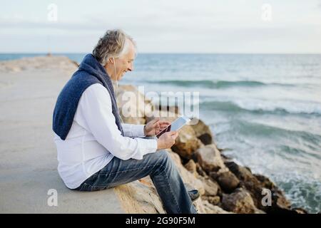 Happy mature man using digital tablet sitting at seaside Stock Photo