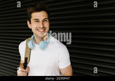 Smiling young man looking away while carrying backpack in front of shutter Stock Photo