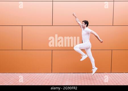 Cheerful young man jumping on footpath by orange wall Stock Photo