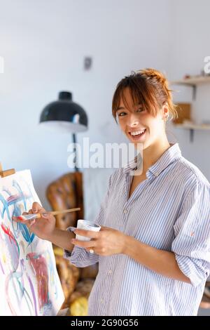 Smiling female painter holding paintbrush while painting in studio Stock Photo