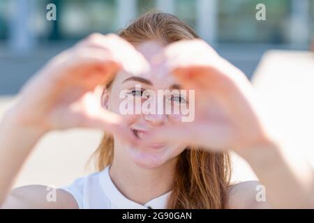 Happy female freelancer looking through heart shape gesture during sunny day Stock Photo