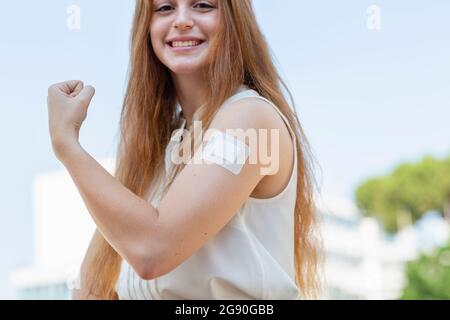Smiling redhead businesswoman flexing muscles after taking vaccine during pandemic Stock Photo