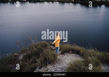 Young woman wearing yellow raincoat standing with arms outstretched at lakeshore Stock Photo