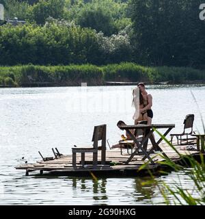 Young couple hugs on the wooden pier on lake in Muzychi in Kiev region, Ukraine on sunny July day their friend sits on the pier his legs in the water. Stock Photo
