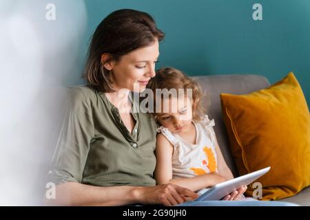 Mother using digital tablet while sitting with daughter at home Stock Photo