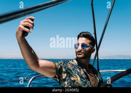 Young man taking selfie through smart phone in yacht on sunny day Stock Photo