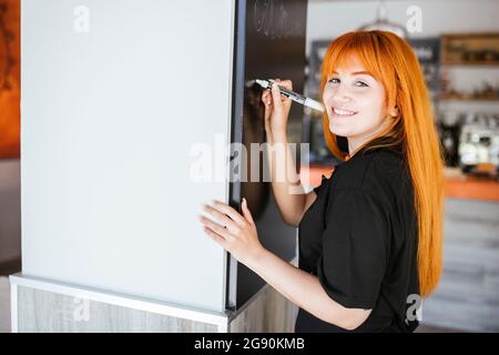 Smiling young redhead waitress writing on blackboard at bar Stock Photo