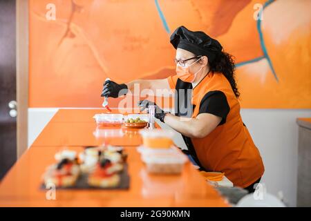 Female chef wearing protective face mask packing food in container at cafeteria Stock Photo