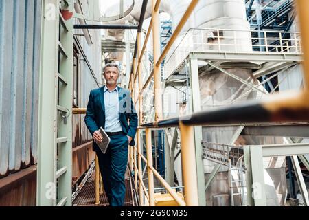 Businessman holding digital tablet while walking with hands in pockets at industry Stock Photo