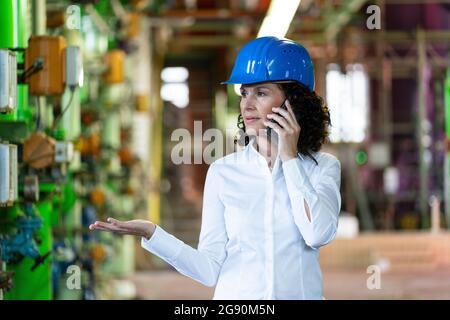 Female engineer with hardhat talking on mobile phone while standing at power station Stock Photo