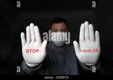 A man in a medical mask and gloves shows STOP with two hands, with the red text STOP COVID 19. on a dark background. Coronavirus infection protection Stock Photo