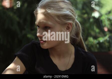 A blond teenage girl in a black T-shirt is sitting outdoors in the greenery and looks away with interest. Portrait of a pretty blonde girl in sunlight. Stock Photo