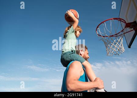 Father and son playing basketball. Sports Concept. Boy child sitting on the dad shoulders, throwing basketball ball into basket, side view on sky back Stock Photo