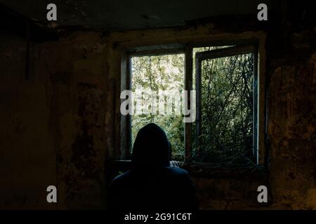 A horror concept. Of a hooded figure, back to camera. Looking out of a broken window. In an abandoned spooky ruined house. Stock Photo