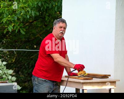 Senior caucasian man with a mustache in a red t-shirt is sanding a board using a sander on a layout in a garden near a white wall of a house Stock Photo