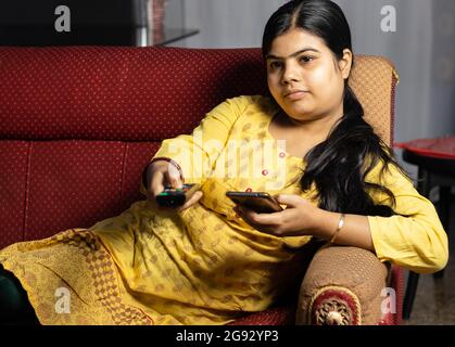 An Indian housewife woman watching television holding remote and mobile phone in hands, sitting on sofa Stock Photo