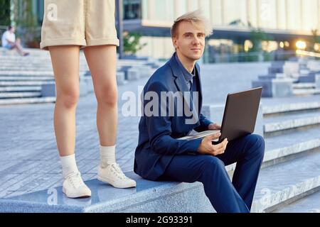 Man with notebook sits on stairs outdoors and woman stands near. Stock Photo