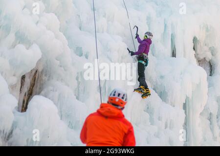 Man standing and controlling a safety top rope while female with ice climbing equipment, climbing on a frozen waterfall Stock Photo