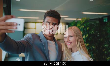 Cheerful black man with laughing blond woman taking selfie with smartphone in modern office having fun. Stock Photo