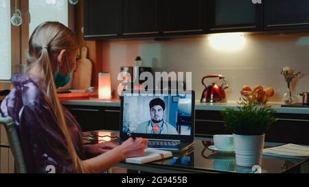 Medium shot of Business woman making video call to a doctor and getting medical consultation from home. She wearing home bathrobe and medical mask. Stock Photo