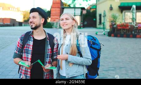 Trendy couple of tourists finding necessary destination on map and admiring surroundings. They standing on city square. Stock Photo