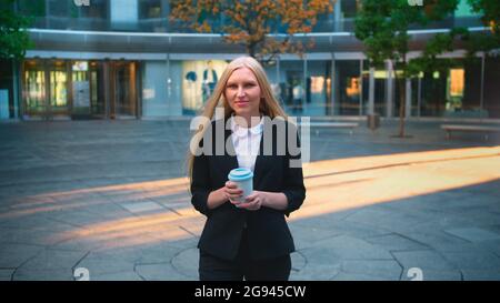 Elegant blond woman in suit and with long hair drinking coffee from white cup in patio with trees. Stock Photo
