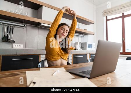 Smiling young brunette woman working on laptop computer while sitting in the kitchen, stretching hands Stock Photo