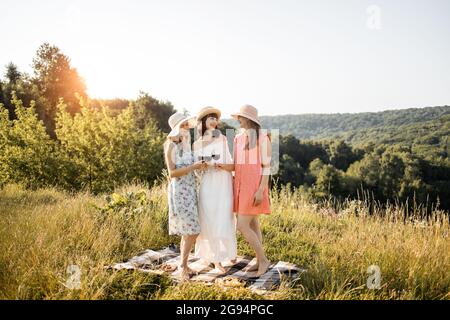 Three beautiful women best friends clinking glasses of red wine, while standing on the grass having cozy picnic time at summer meadow on sunny day Stock Photo