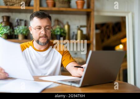 A middle-aged man works with papers at home in front of a laptop monitor. He is sitting with glasses and typing text. Modern technology concept. Stock Photo