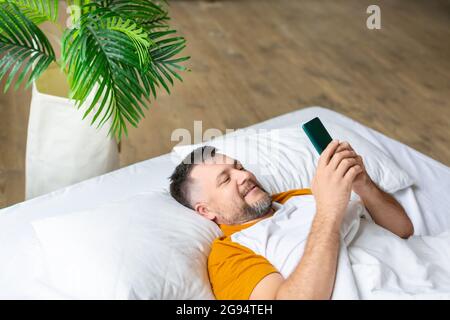 Middle aged man in bed checking his messages in smartphone after awakening. He lying in bed and smiling. Addiction to social networks concept. Stock Photo