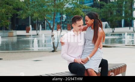 Two mixed-race lovers sitting on a bench and kissing. They having a date in a beautiful place in the city center. Stock Photo