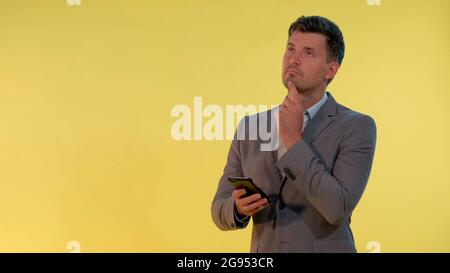 Young businessman in suit looking on smartphone and thinking about something on yellow background. He probably making some plans. Stock Photo
