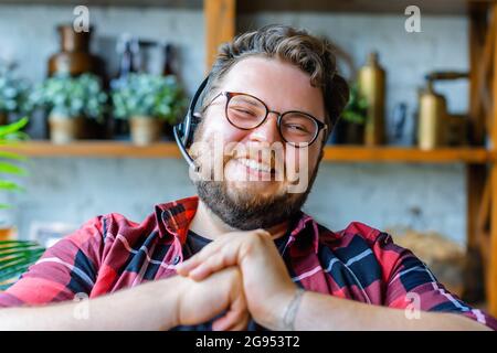 Happy laughing young man in headset during online video call. He looking at camera and smiling. Online meeting with friends or relatives. Stock Photo