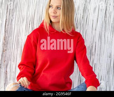 Smiling blond woman wearing red blouse or hoodie sits in front of camera. On her hoodie in place for christmas inscription. Winter outfit with copy space for mockup. Stock Photo
