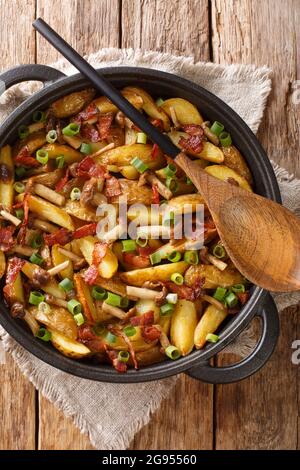 Fried potatoes with bacon and mushrooms close-up in a frying pan on the table. Vertical top view from above Stock Photo