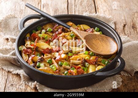 Freshly fried potato wedges with green onions, mushrooms and bacon close-up in a frying pan on a wooden background. horizontal Stock Photo