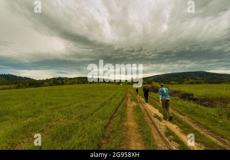 Krakow, Poland - April 10, 2021: Three unidentified people with backpack hike or trek on a trail in the middle of a field in countryside on a way to l Stock Photo