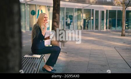 Side view of formal woman sitting with tablet on bench and drinking coffee looking pensively away in leisure Stock Photo