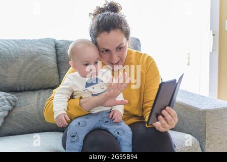 young woman with her baby making a video call with her mobile phone in her living room. Family time Stock Photo