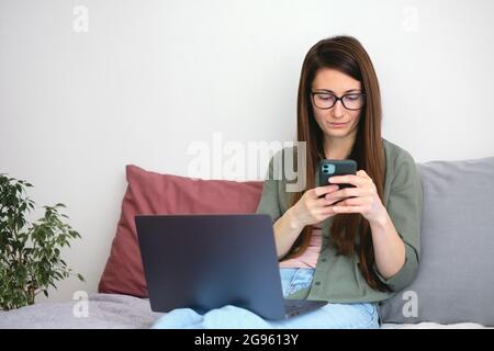 Young woman freelancer typing message, sitting in bed and working with laptop and smartphone. Life at home. Slow life concept. Stock Photo