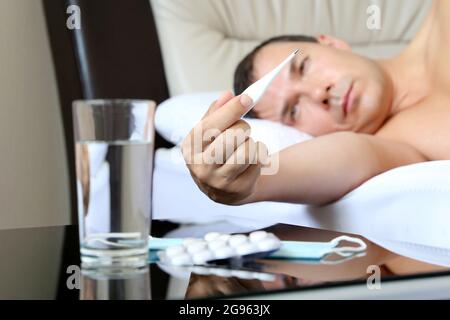 Sick man lying with digital thermometer in a bed, pills and water glass in the foreground. Concept of illness, fever, coronavirus symptoms Stock Photo
