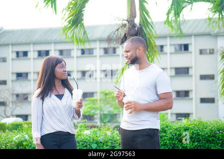 African friends consisting of a guy and a lady joyfully interacting together outdoor as they talk and smile with refreshing drinks in their hands Stock Photo