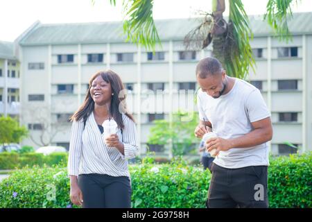 African friends consisting of a guy and a lady joyfully interacting together outdoor as they talk and smile with refreshing drinks in their hands Stock Photo