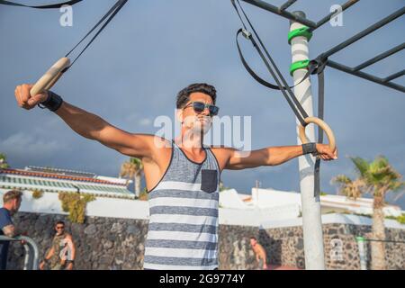 Athlete trains with rings in an outdoor gym. Man with sunglasses performs gymnastic rings workout - concept of sporty person training lifestyle Stock Photo