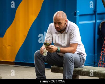Medellin, Colombia - July 21 2021: Bald Latin Men in White Shirt and Blue Jeans Sits on a Bench and Looks at his Phone Stock Photo