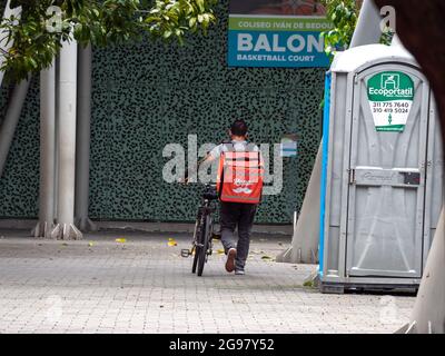 Medellin, Colombia - July 21 2021: Latin Men Walks Beside his Bicycle with 'Rappi' Backpack in a Park with a Sign that Reads : 'Balon' Stock Photo