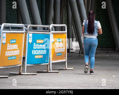 Medellin, Colombia - July 21 2021: Latin Woman Walks by a Waiting Line for Vaccination with Banners that Read: 'In Medellin, We're Vaccinating Against Stock Photo