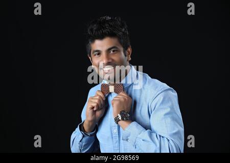 Close up portrait - Indian man ties a bowtie at the collar, correcting wood bow on his grey background Stock Photo