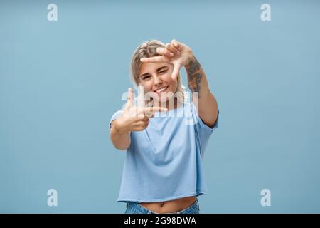 Be creative and happy. Portrait of charming enthusiastic female designer with blond hair and tattoo on arm winking and smiling joyfully making frame Stock Photo